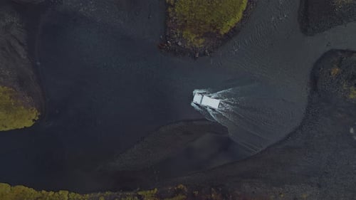 Top-down aerial view of white off-road car going across the water on a black gravel road.