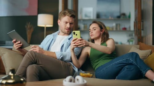 Serious Woman Showing Mobile Phone To Boyfriend Indoors. Involved Spouses Reading Devices At Livi...