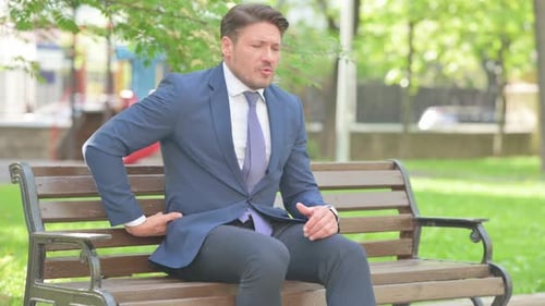 Man in Suit Sits with Back Pain Outdoors