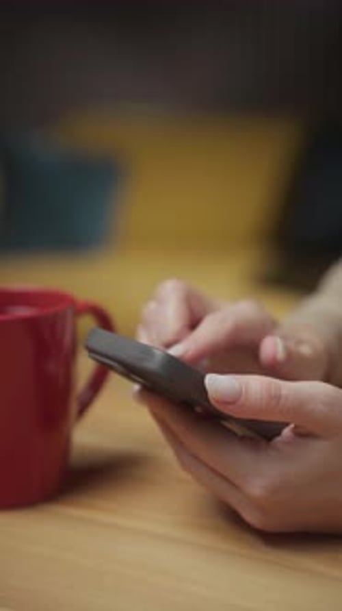 Person Using Smartphone While Sitting at a Table with a Red Mug During a Cozy Indoor Setting