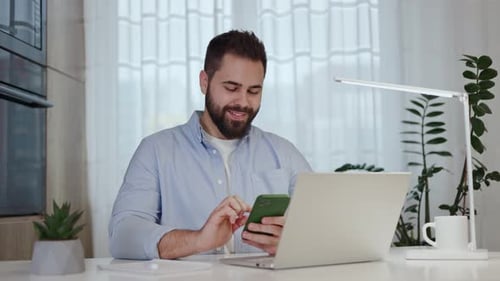 Man Using Phone and Laptop at Desk Indoors