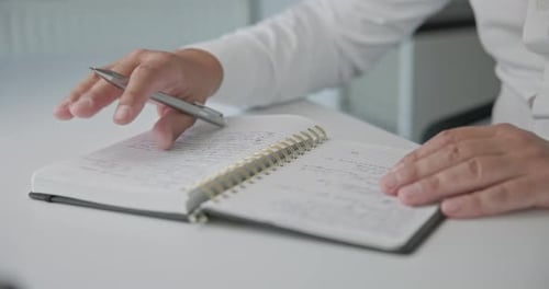 A Closeup of a Man in a Shirt Checking Notes and Making Corrections with a Pen in a Notepad on a
