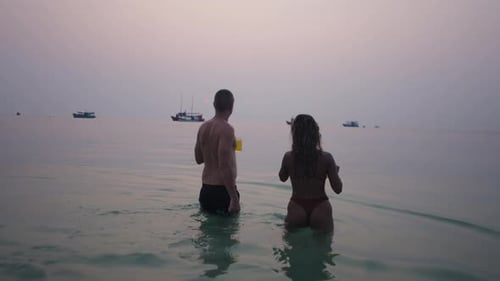 Couple Walks into Ocean at Sunset with Drinks