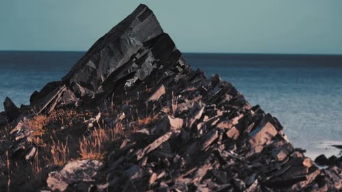 A closeup of dark sharp rocks formations with patches of dry grass and lichen. Dark blue sea in the