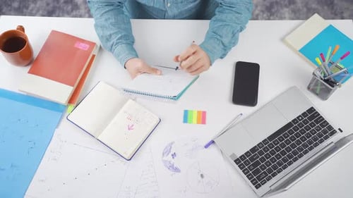 Person Writing at Desk with Papers and Technology