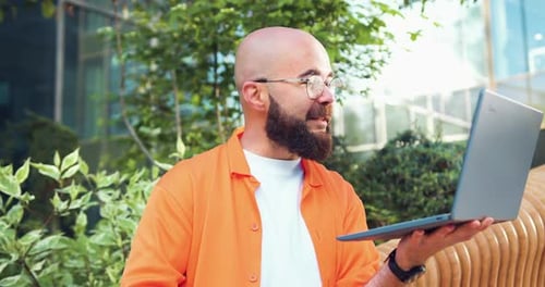 Apuesto hombre hipster con gafas sentado afuera en el banco, trabajando en el portátil. Negocio barbudo