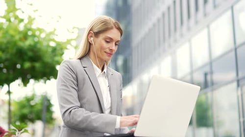 Businesswoman works on a laptop sitting on a bench on street near an office building. Female