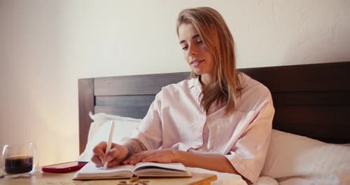 A Blonde Girl in a Light Pink Nightgown Makes Entries in Her Diary Early in the Morning with a Cup
