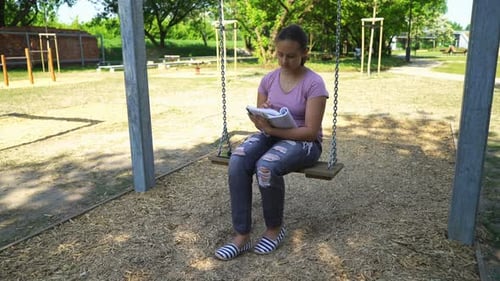 Teenage girl reads book on a swing enjoying a summer day at the city park