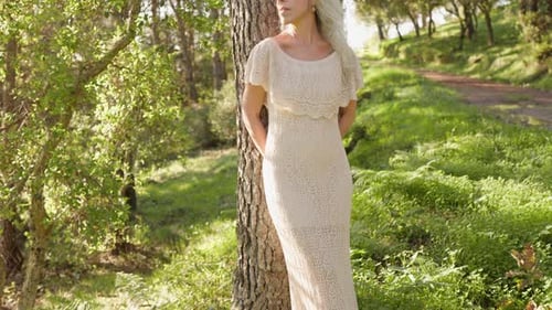 Serene Senior Woman in Cream Dress Leans Against Tree in Lush Green Forest