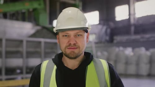 Man Stands at a Factory Wearing a White Helmet of Heavy Industry Engineer and a Protective Uniform