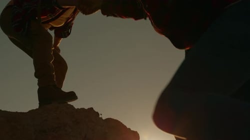 Silhouette woman lends helping hand to companion while climbing to top of mountain in travel