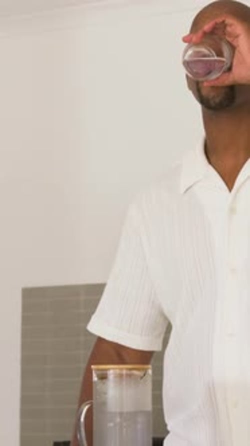 Vertical video: Lifting glass, man in white shirt sipping, placing glass beside pitcher in kitchen