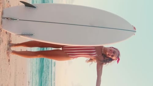 Young Cheerful Caucasian Woman Showing Shaka Sign and Surfboard Stands on Beach