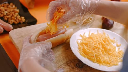 Hands Add Shredded Cheese to Hot Dog