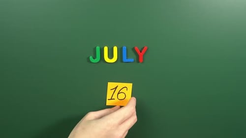 Hand sticking a sticker 16 July calendar day on school board. 16 date of July. Sixteenth day