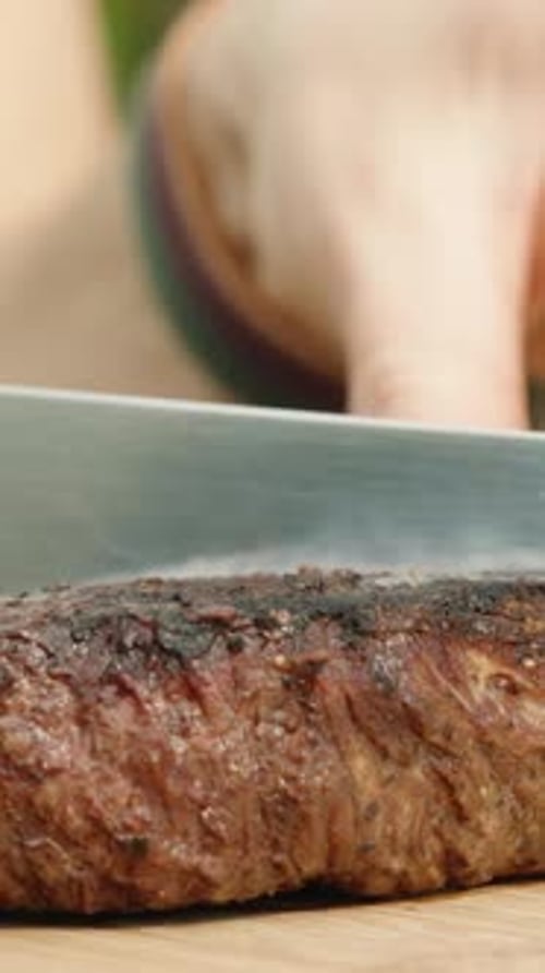 Vertical. I cut a piece of beef steak in half with a knife on a wooden cutting board, close-up.