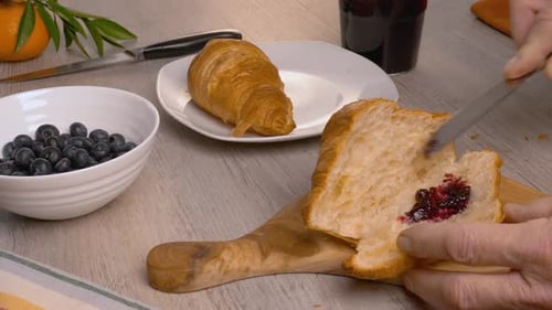 A man puts blueberry jam into croissants with a knife on a wooden cutting board.