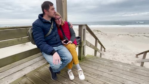 Loving Couple Sits on a Wooden Bench By the Beach on a Cloudy Day They Cuddle Kiss and Talk