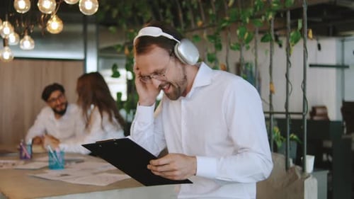 A Businessman Wearing Headphones is Smiling and Dancing in a Busy Office Colleagues are Working