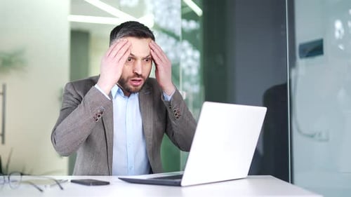 Frustrated stressed businessman reads bad news on a laptop while sitting at workplace in business