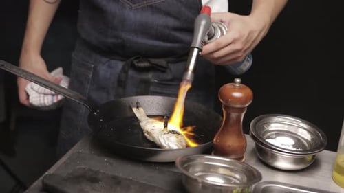 Chef Using Butane Gas Torch To Scorch Fresh Fish In A Cast-iron Pan. close up