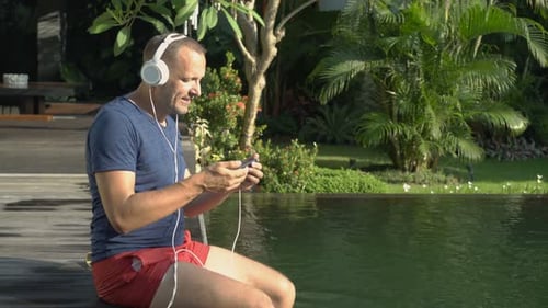Handsome young man enjoying a movie on his smartphone by the tropical swimming pool