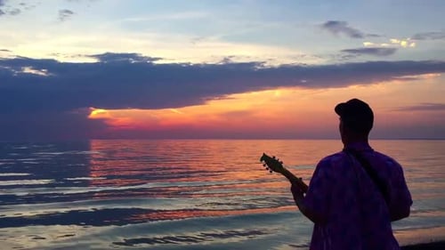 Handheld on man with back to camera playing a busker toy guitar to the setting sun on a beautiful ev