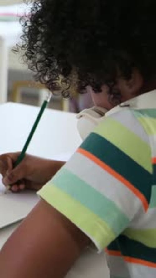 Vertical video: Reaching for green pencil, striped-polo teen at home table, working on homework
