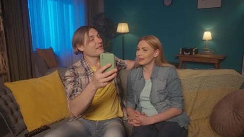 A Couple is Talking on a Video Call on a Smartphone While Sitting on the Couch Couple Waving in