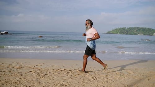 Grey Bearded Elderly Man Runs with Modern Fitness Tracker Watch in Morning on Ocean Beach Listens to