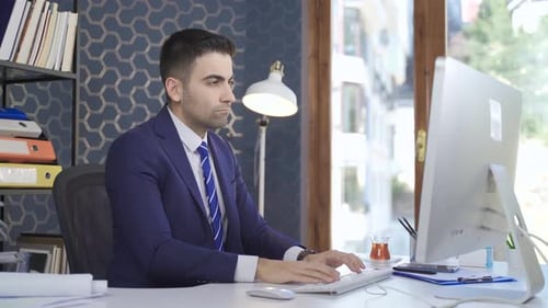 Thoughtful Looking Office Worker Man Looking At Computer While Sitting At Desk.