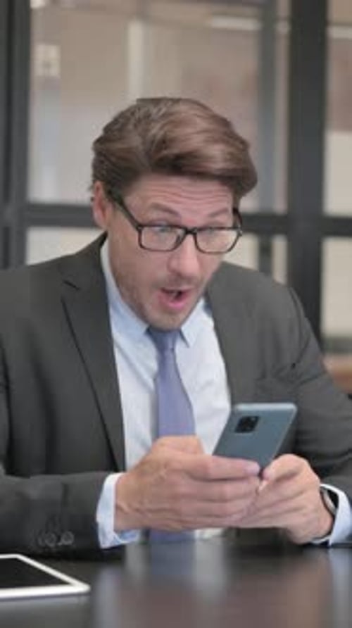 Businessman Celebrating Online Success on Phone while Sitting in Office, vertical