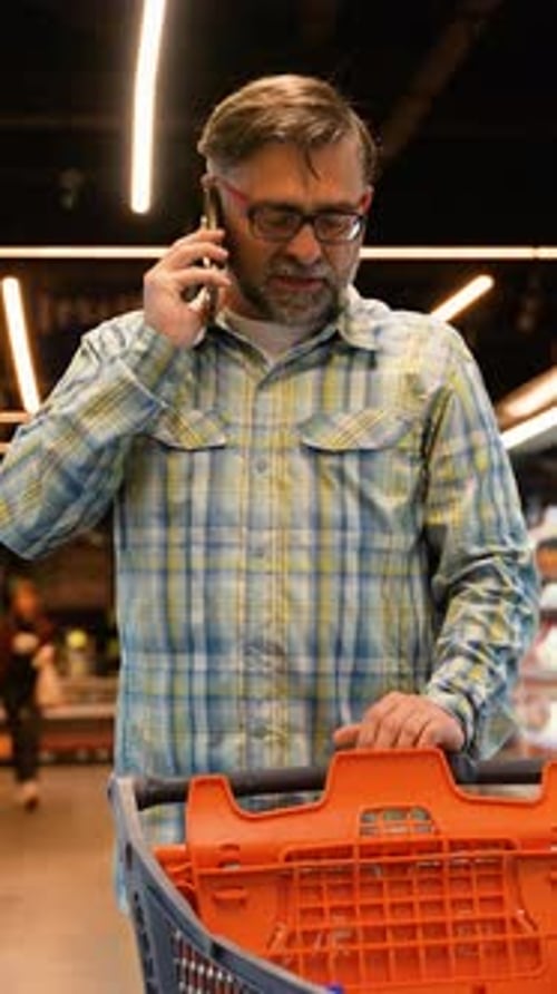 A Man 50s with a Trolley in a Supermarket Talks on the Phone with His Wife Asking What to Buy