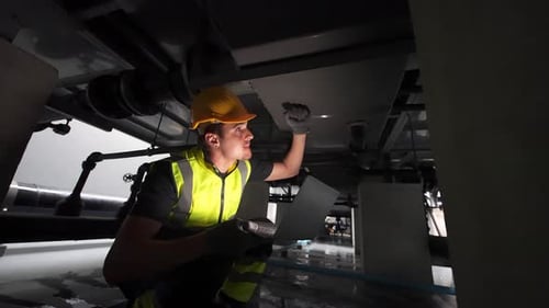 Worker inspects machinery under facility with laptop during maintenance operation