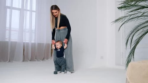 Mother teaching her baby to walk. Long-haired woman holds an infant by the hands. Room backdrop.