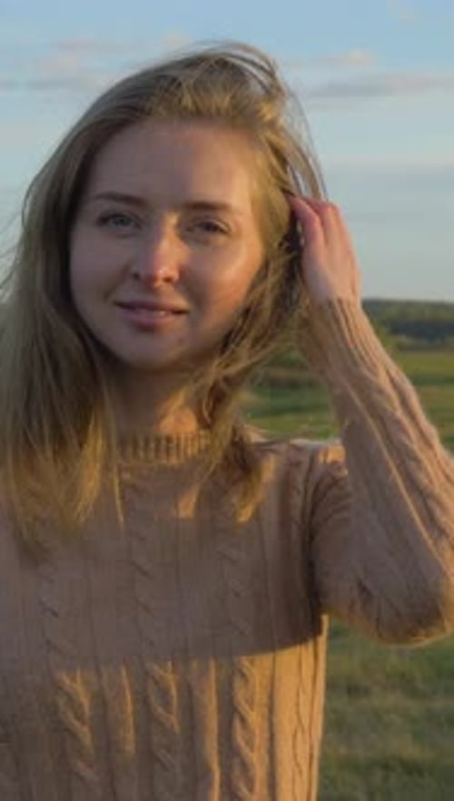 Portrait of Young Woman Smiling and Touching Her Hair at Sunny Evening in Green Field Medium Shot