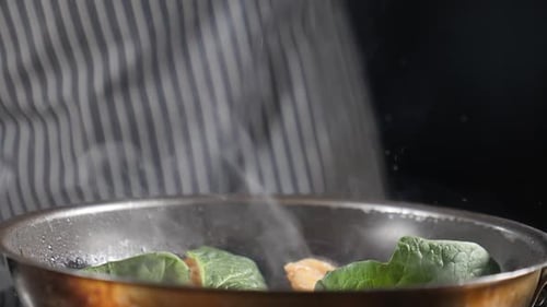 Le chef vêtu d'un tablier rayé ajoute des feuilles d'épinards fraîches dans une poêle chaude, de la vapeur s'échappe de la poêle