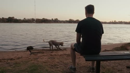 Man sits with his dog on the small bench by the lake