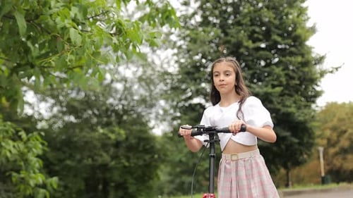 A Little Beautiful Girl with Dark Hair is Riding an Electric Scooter in the Park on a Sunny Day