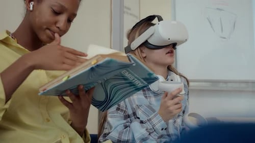 Black Girl Reading Book near Female Classmate Equipped with VR Headset
