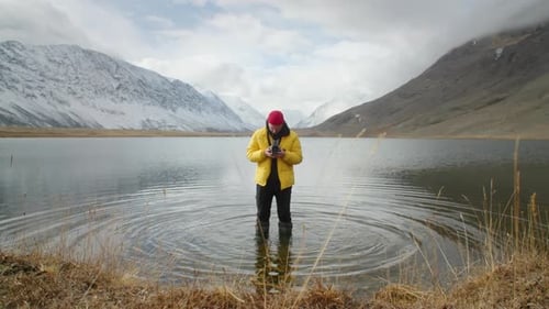 Young Guy Adventurer Dressed in Bright Yellow Jacket Standing in Serene Mountain Lake Holds an Old