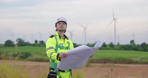 Engineer holding blueprint and looking at wind turbines, standing in renewable energy field,