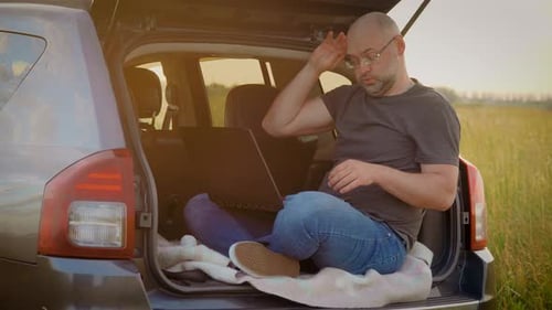 Homem de negócios cansado trabalha duro em um laptop respirando profundamente sentado no porta-malas do carro enquanto