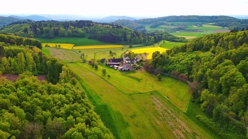 A Farm Stands on a Hill with Crop Fields White Bales of Hay and Pastures Surrounded By Forest