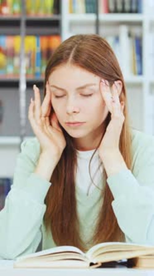 Exhausted Female Student Holding Head with Eyes Closed While Studying in Library