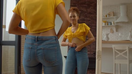 Overweight Black Woman Trying to Button Up Tight Pants in Front of Mirror
