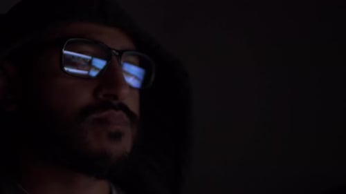 Homme asiatique à capuche avec reflet d'écrans d'ordinateur vus dans des lunettes dans une pièce sombre. Verrouillé