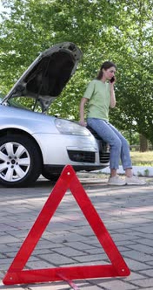 Worried woman talking on smartphone near broken car and warning triangle outdoors