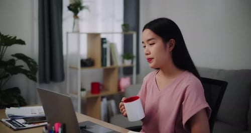 Junge Frau hält Kaffee zum Trinken bereit, während sie zu Hause am Laptop arbeitet.